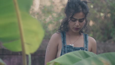 Toma-Mediana-De-Mano-De-Una-Joven-Y-Bonita-Modelo-India-Parada-En-El-Jardín-Rodeada-De-Plantas-Vestida-Con-Un-Vestido-De-Mezclilla-Con-Una-Mirada-Seria-Hacia-Abajo