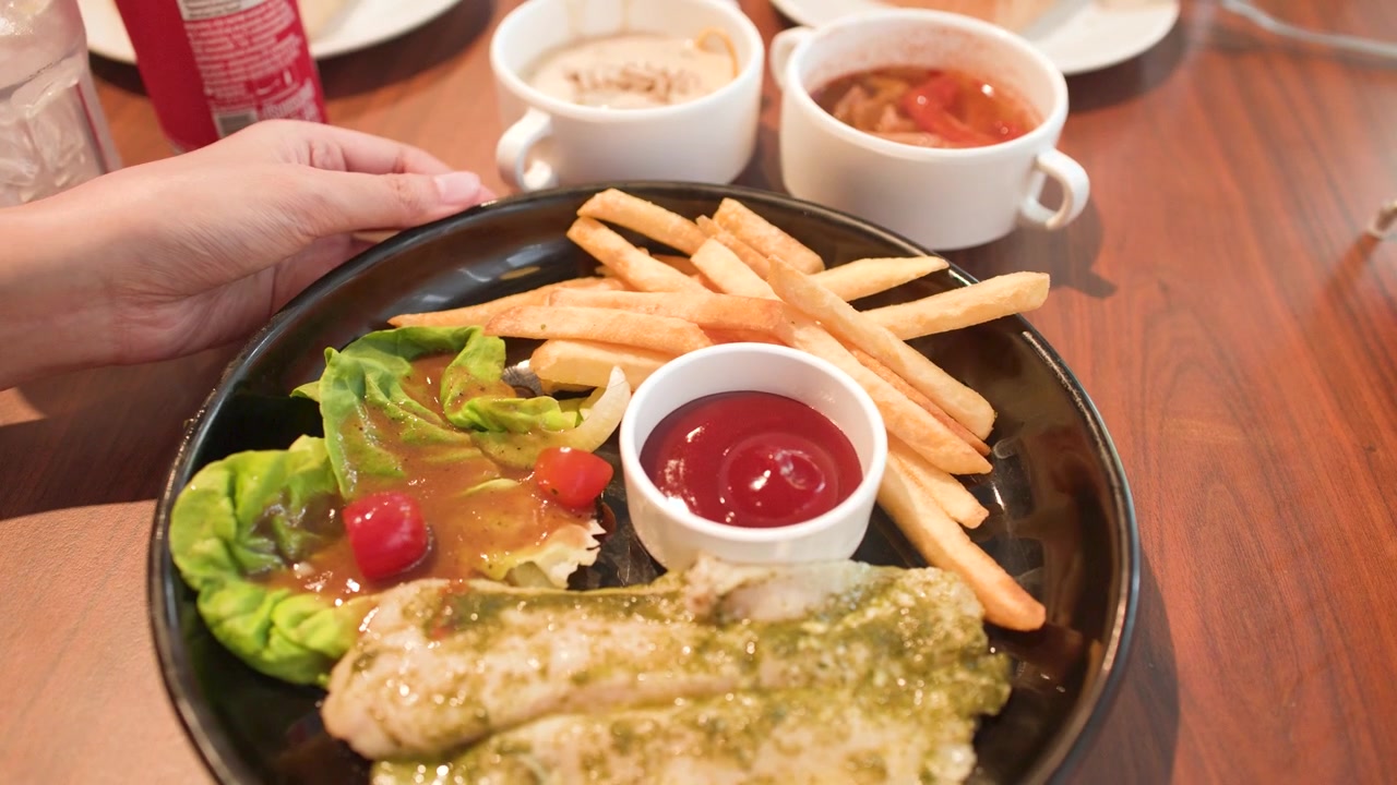 Una persona ruota un piatto nero contenente pesce alla griglia, patatine fritte e salse d'accompagnamento.