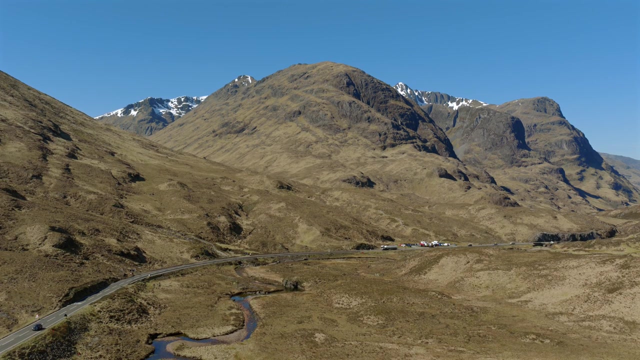 Static drone shot of traffic flowing through Glencoe in the Scottish Highlands. Shot in April with snow still visible on the high level peaks. The road is the A82 from Tyndrum to Fort William.