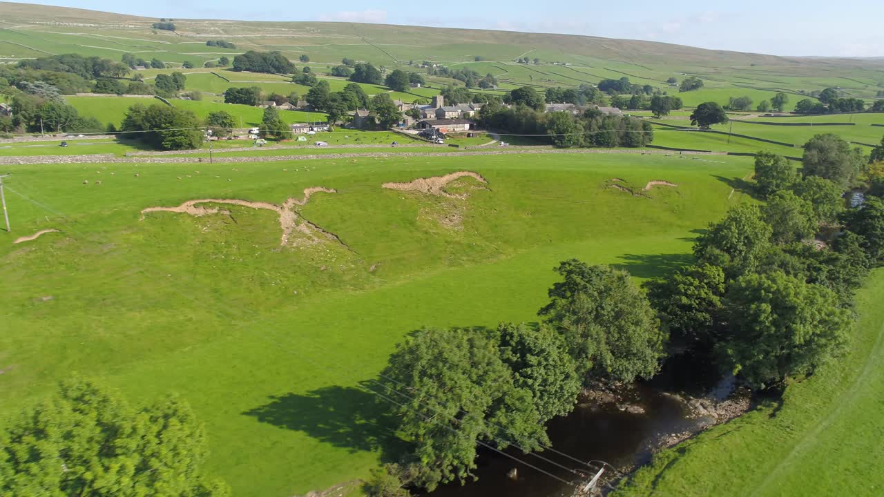 imágenes de drones volando paralelas a un río bordeado de árboles en la zona rural de yorkshire cerca de ingleton en un soleado día de verano, rodeados de campos, el campo y un pequeño pueblo con colinas en el fondo