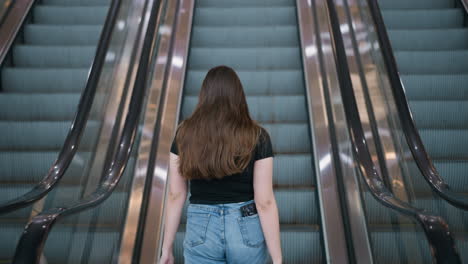 back view of young woman with long brown hair wearing black t-shirt and blue jeans stepping onto escalator in modern shopping mall holding shopping bags in both hands under bright lighting