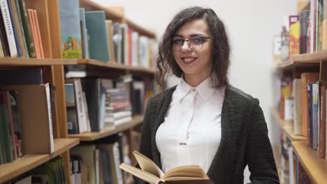 friendly teacher holds a book in the library and smiles at the camera