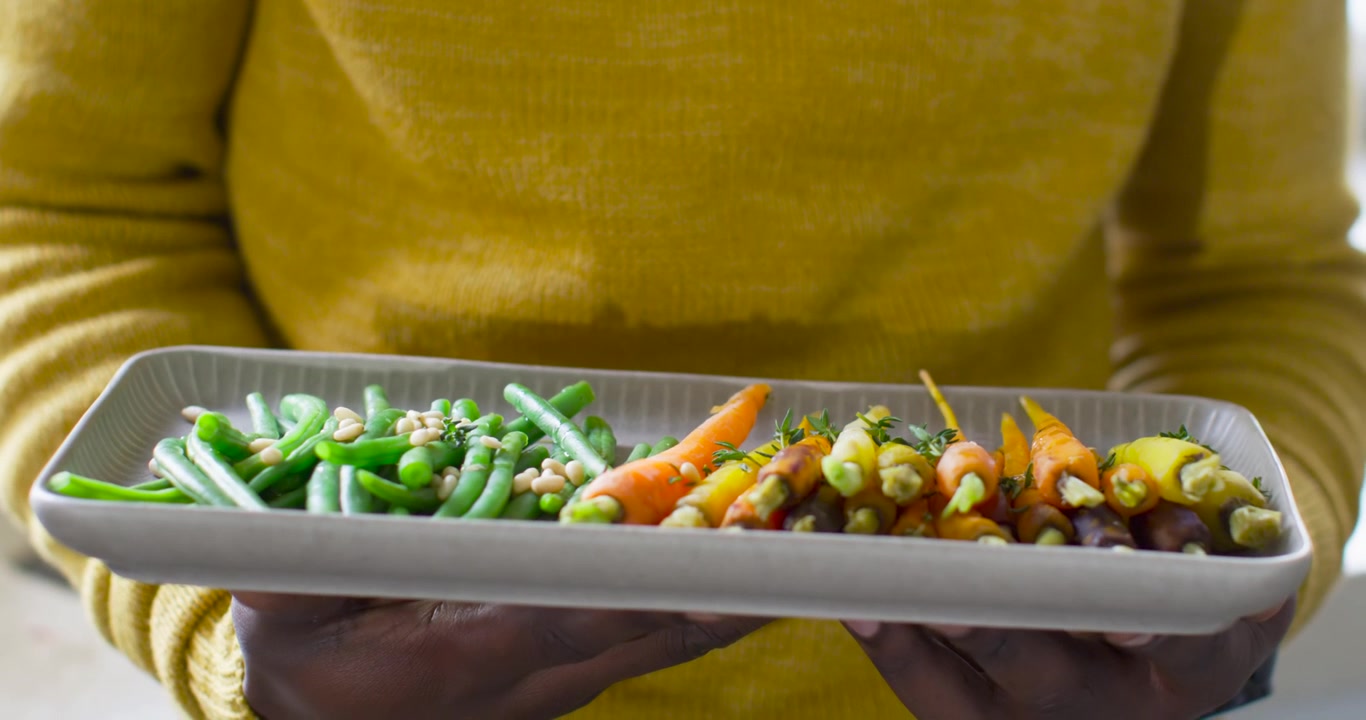 Personne en pull moutarde abaissant un plat après un léger mouvement de la main pour présenter des légumes près de la fenêtre