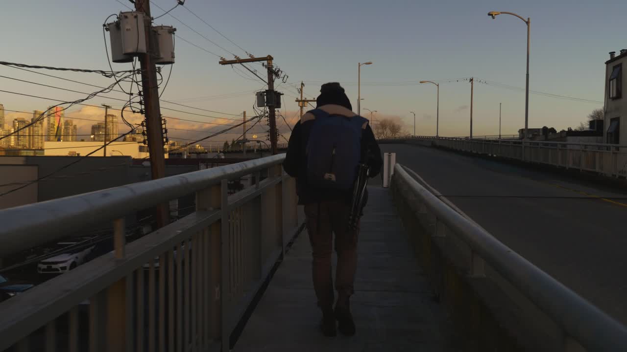Seguendo un viaggiatore che cammina lungo il marciapiede con guardrail su un ponte in città, mentre le auto passano di sera con le montagne sullo sfondo, Vancouver BC.