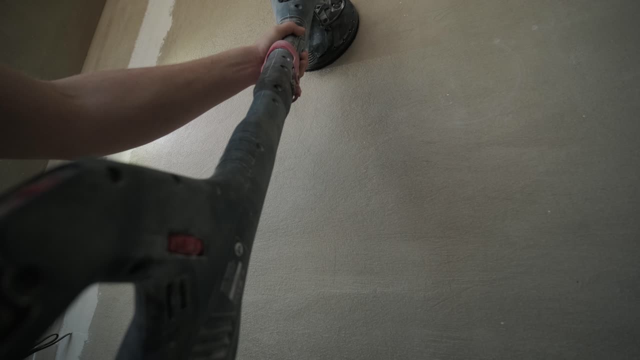 Close-up image of a hand holding a power sander against a freshly plastered wall, focusing on the tool and surface texture