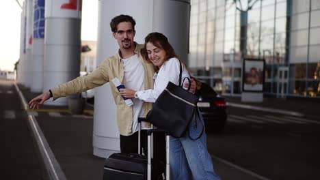 Pareja-Joven-Y-Elegante-Con-Equipaje-Parado-En-El-Estacionamiento-Del-Aeropuerto-Con-Sus-Maletas.-Esperando-El-Coche-Después-De-La-Llegada.-Hombre-Intentando