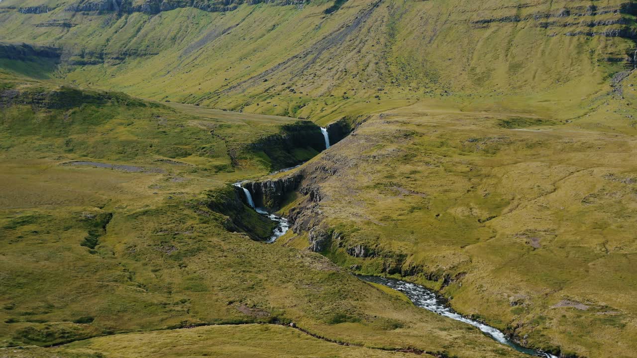 vuelo de avión no tripulado sobre una cascada que desciende en medio de las colinas verdes en islandia, en verano.