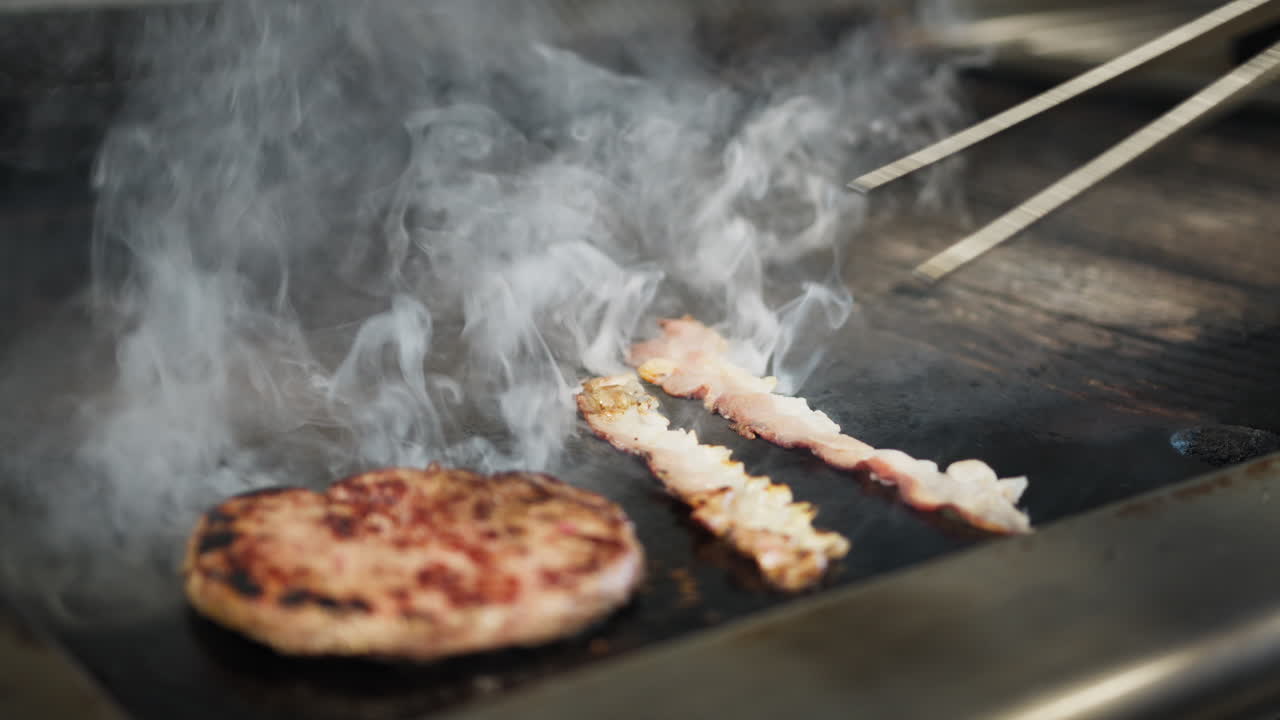 Chef Flipping The Bacon On The Professional Kitchen Griddle With The Tongs