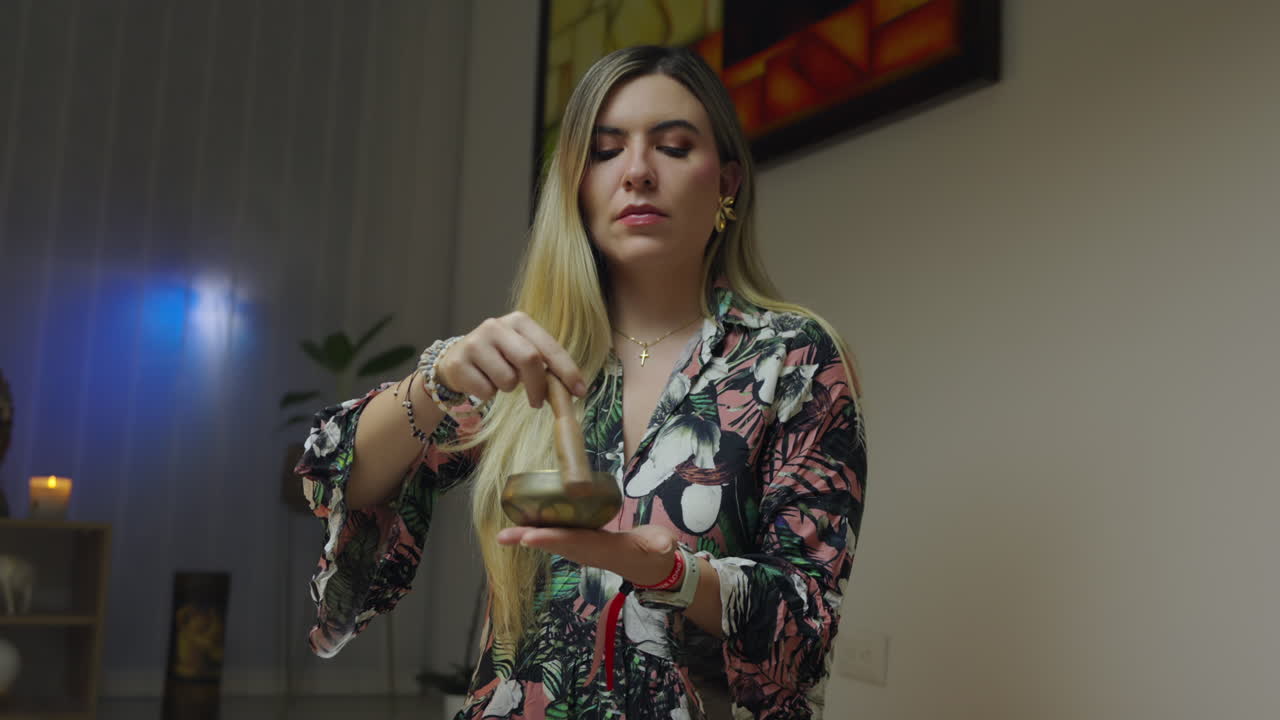 Blonde woman uses Tibetan singing bowl indoors for relaxation at night