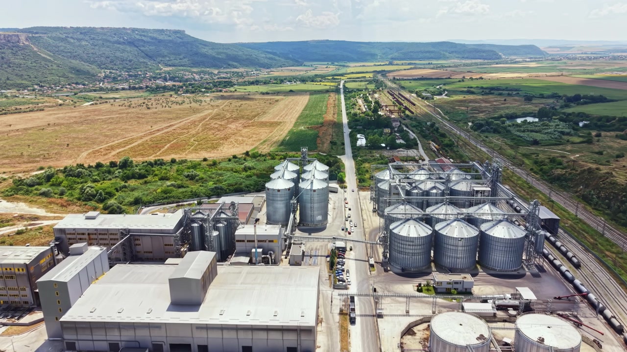 Große Industrieanlage mit Silos und einer Bahnstrecke in der Nähe eines Feldes und Hügeln unter klarem Himmel