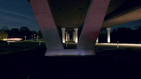 Timelapse-under-a-bridge-with-moving-cars-creating-light-streaks-during-traffic-in-a-blue-cool-night-with-a-view-of-trees-and-the-sky-in-the-background