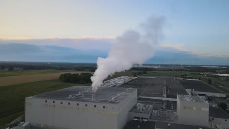 órbita-Aérea-De-Una-Fábrica-Con-Vapor-Saliendo-De-La-Tubería,-Con-Una-Hermosa-Puesta-De-Sol-En-El-Fondo