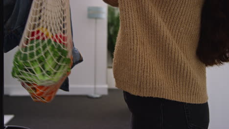 close up of woman getting home from food shopping hanging up coat and carrying bag on fresh vegetables indoors into kitchen