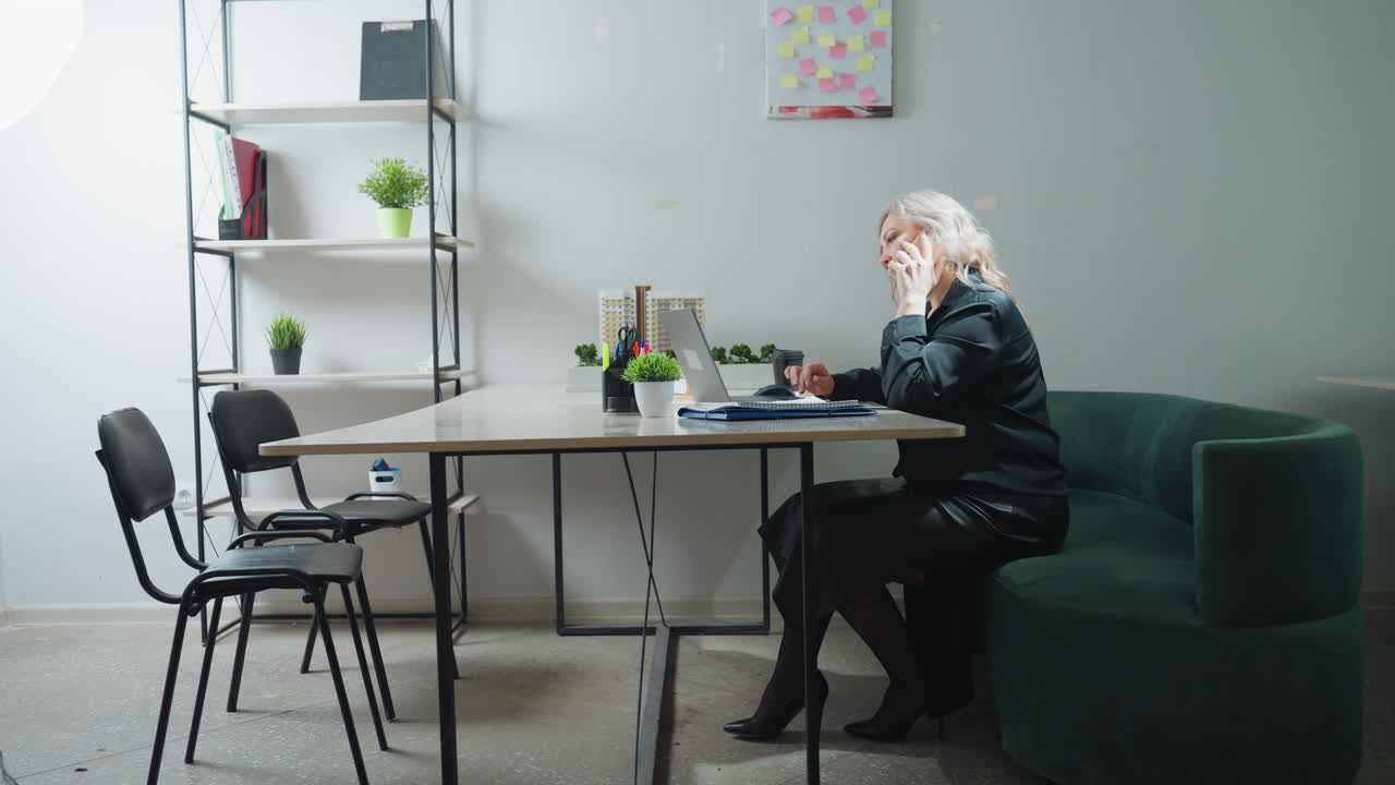 Realtor seated on green couch in bright modern office picks up phone call while multitasking with laptop, surrounded by real estate model, organized desk supplies, and minimalist shelving decor