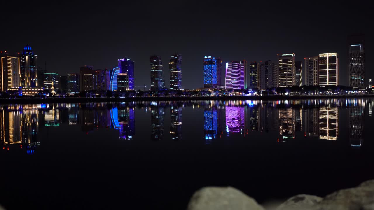 En vidstrakt panoramautsikt over den strålende opplyste bysilhuetten i Lusail, som kaster en perfekt, fargerik refleksjon på det rolige vannet i marinaen om natten.