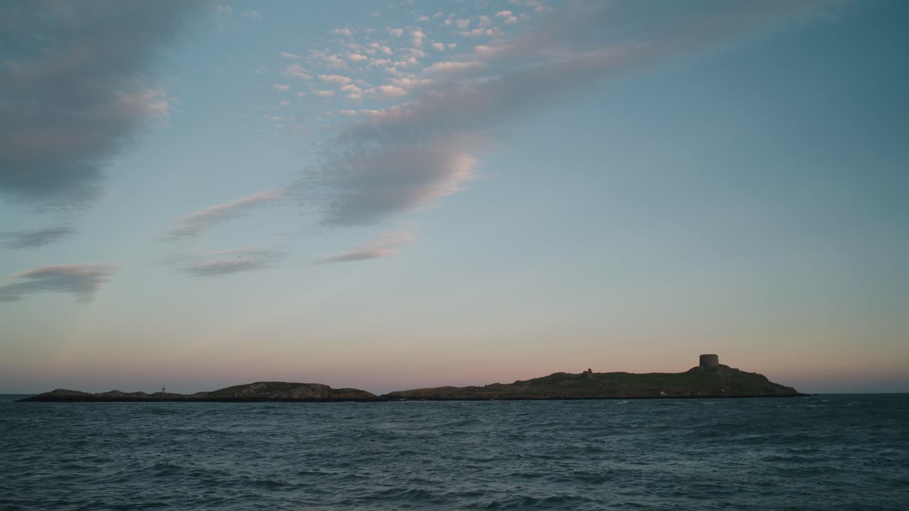 A locked-off shot of Dalkey Island in Dublin, Ireland, with a Martello Tower. The sea ripples in the foreground under a pastel sunset sky with scattered clouds.