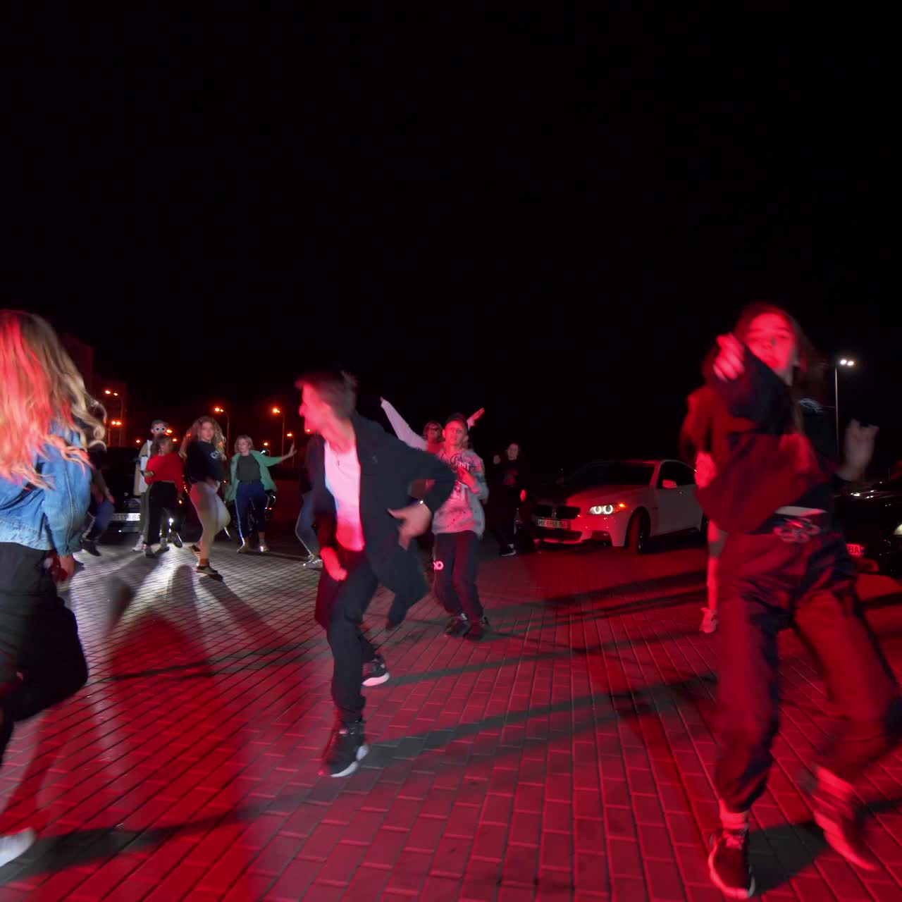Young active people performing hip hop outdoors in the evening. Party in the street. Race cars with headlights on