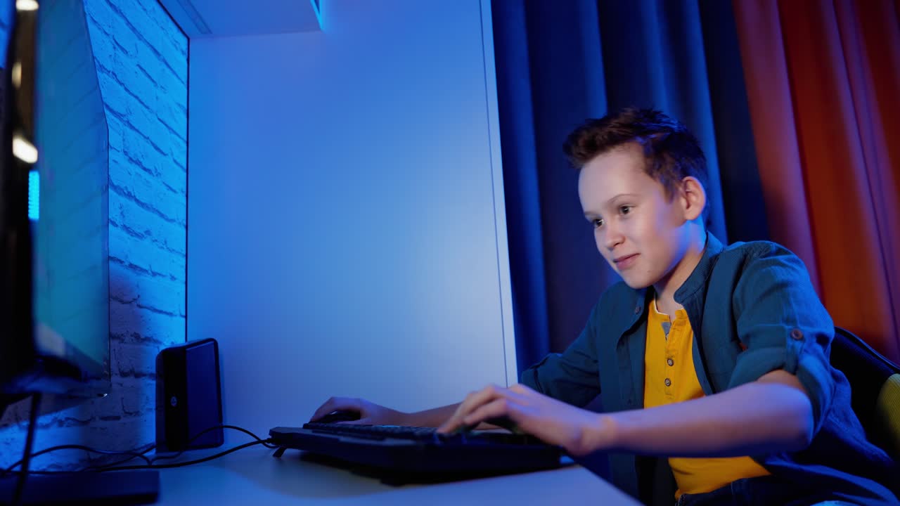 Portrait of teenager playing games. Happy boy sitting in his room and pushing the keyboard buttons while playing on PC. Blue evening light.