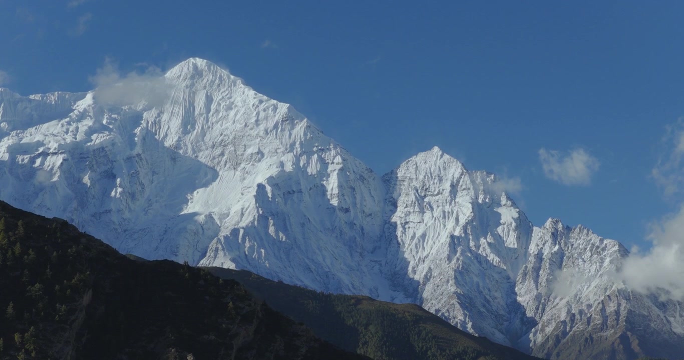 Un'inquadratura aerea ascendente inizia in basso contro il pendio di una collina verde vibrante prima di salire più in alto per rivelare la vista delle montagne Nilgiri innevate sotto un cielo azzurro limpido nel Mustang, Nepal.