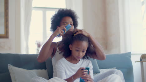 cute black daughter playing mobile game on her smartphone sitting on bed while mum combing her hair