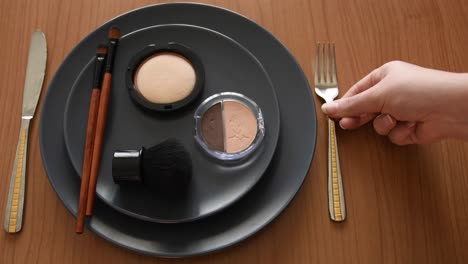 Fondo-De-Concepto-Mínimo-Con-Una-Mano-Femenina-Blanca-Poniendo-Un-Tenedor-En-Un-Lado-De-Un-Plato-Gris-Con-Maquillaje,-Cepillos-Y-Polvos-En-él,-En-El-Otro-Lado-Hay-Un-Cuchillo,-En-Una-Mesa-De-Madera