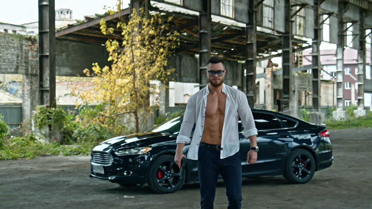 Handsome man with a weapon in abandoned place. Sporty guy in stylish glasses and white unbuttoned shirt with a pistol in his hand on black car background.