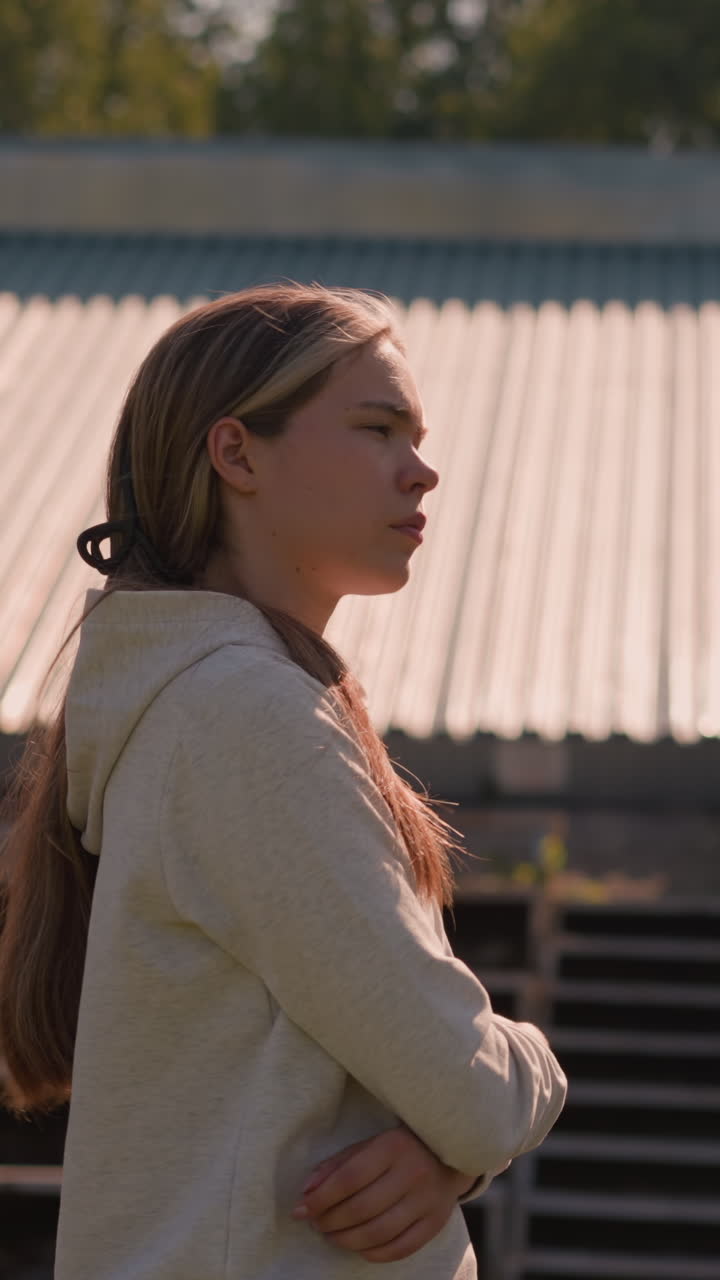 mujer con el cabello largo atado hacia atrás, con una sudadera con capucha, camina por las gradas vacías del estadio en un día soleado, brazos cruzados, parece profundamente en pensamiento, rodeada de asientos vacíos