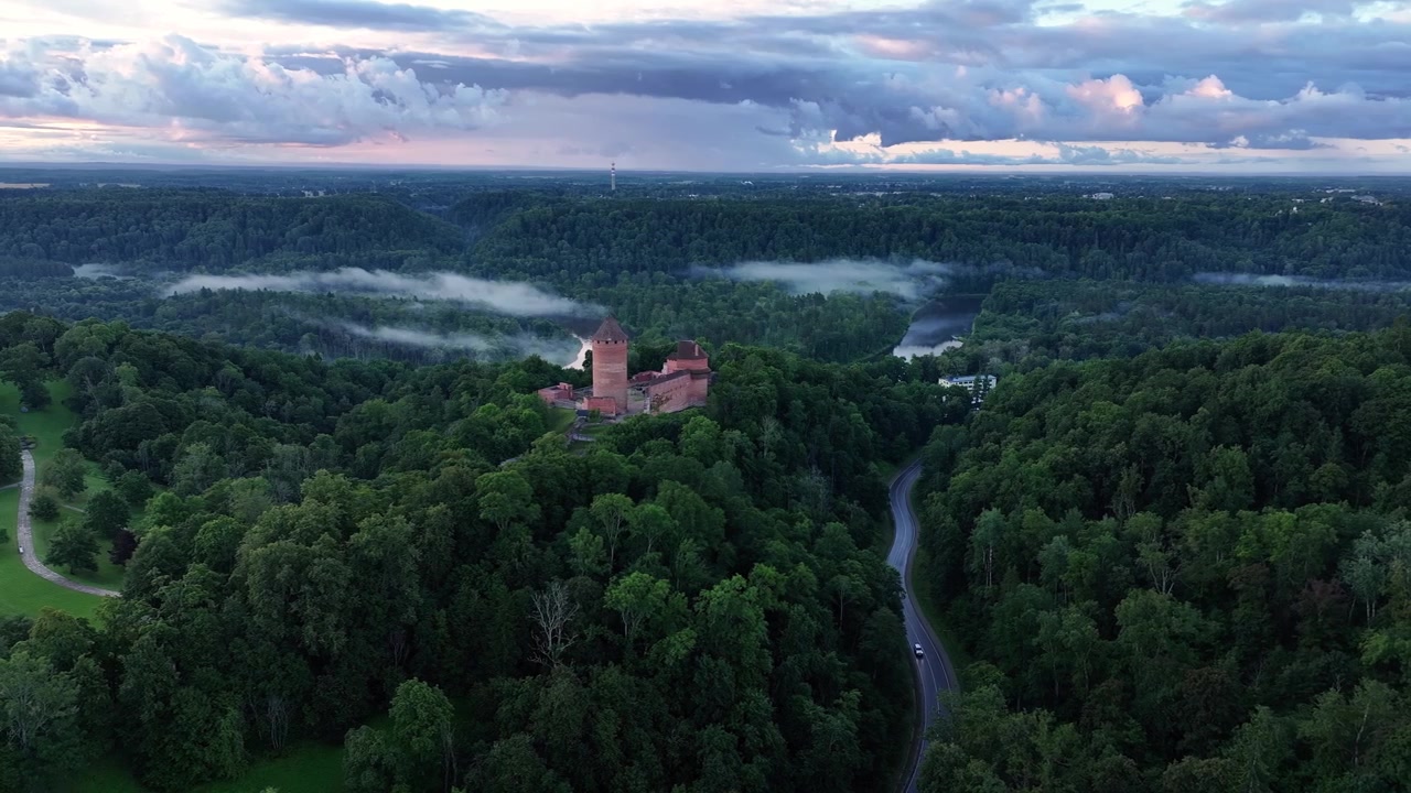 Cinematisk luftdroneudsigt over Turaida Slot omgivet af tætte grønne skove, Gauja-floddalen og lav morgentåge under dramatiske stormskyer i Sigulda, Letland