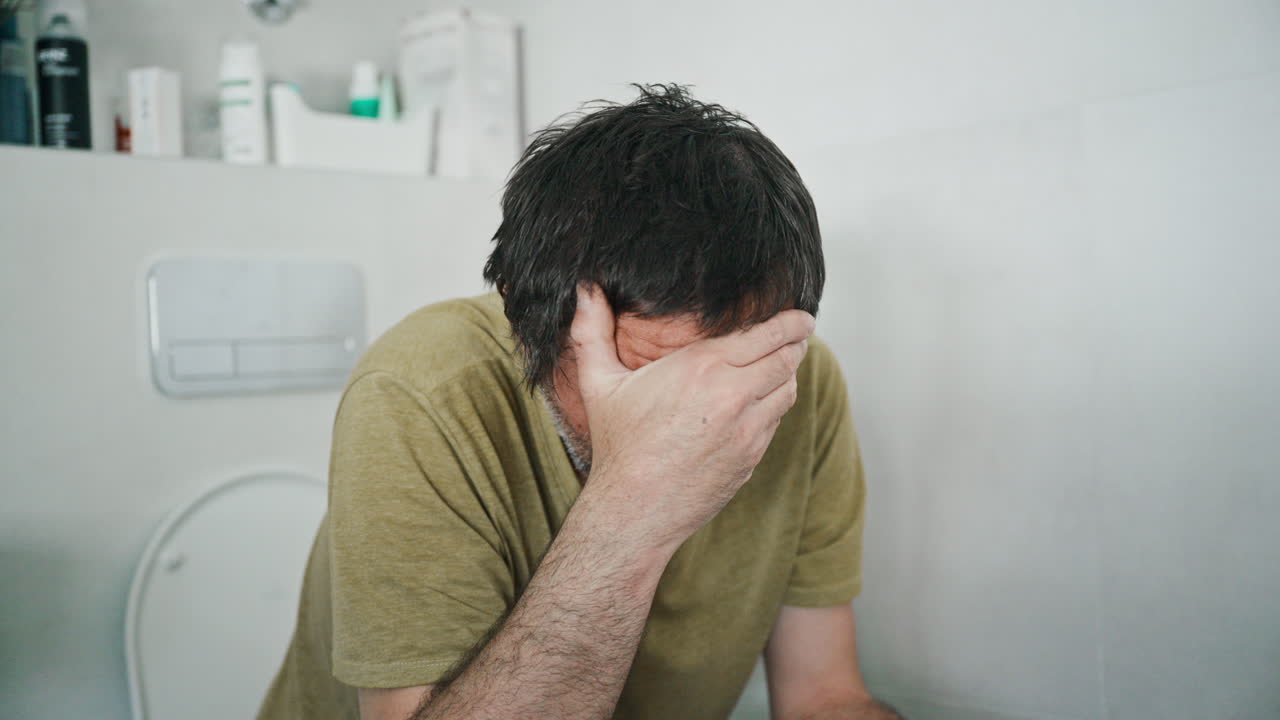Uomo stressato seduto sul water che si copre il viso in bagno, sentendosi male e sopraffatto, 4K.