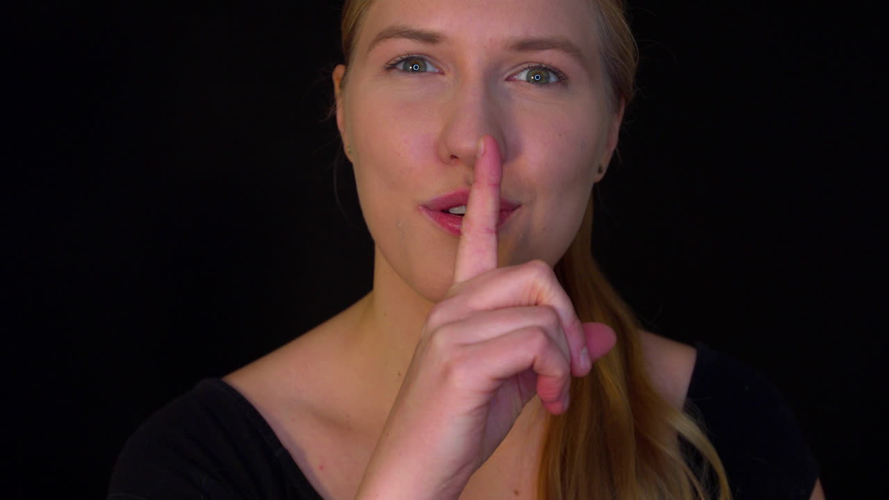 The young woman has a secret. A smiling woman puts a finger to her lips and makes a silence or secret gesture