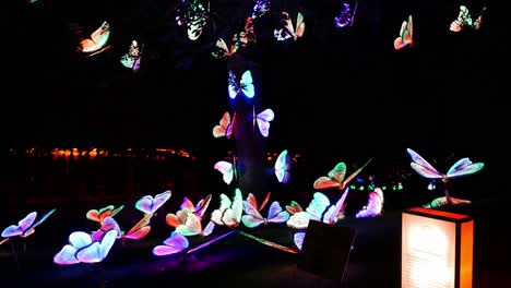 colorful animatronic butterflies sit are scattered on the ground and in a tree while opening and closing their wings