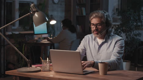 man with headset working on laptop in an office at night