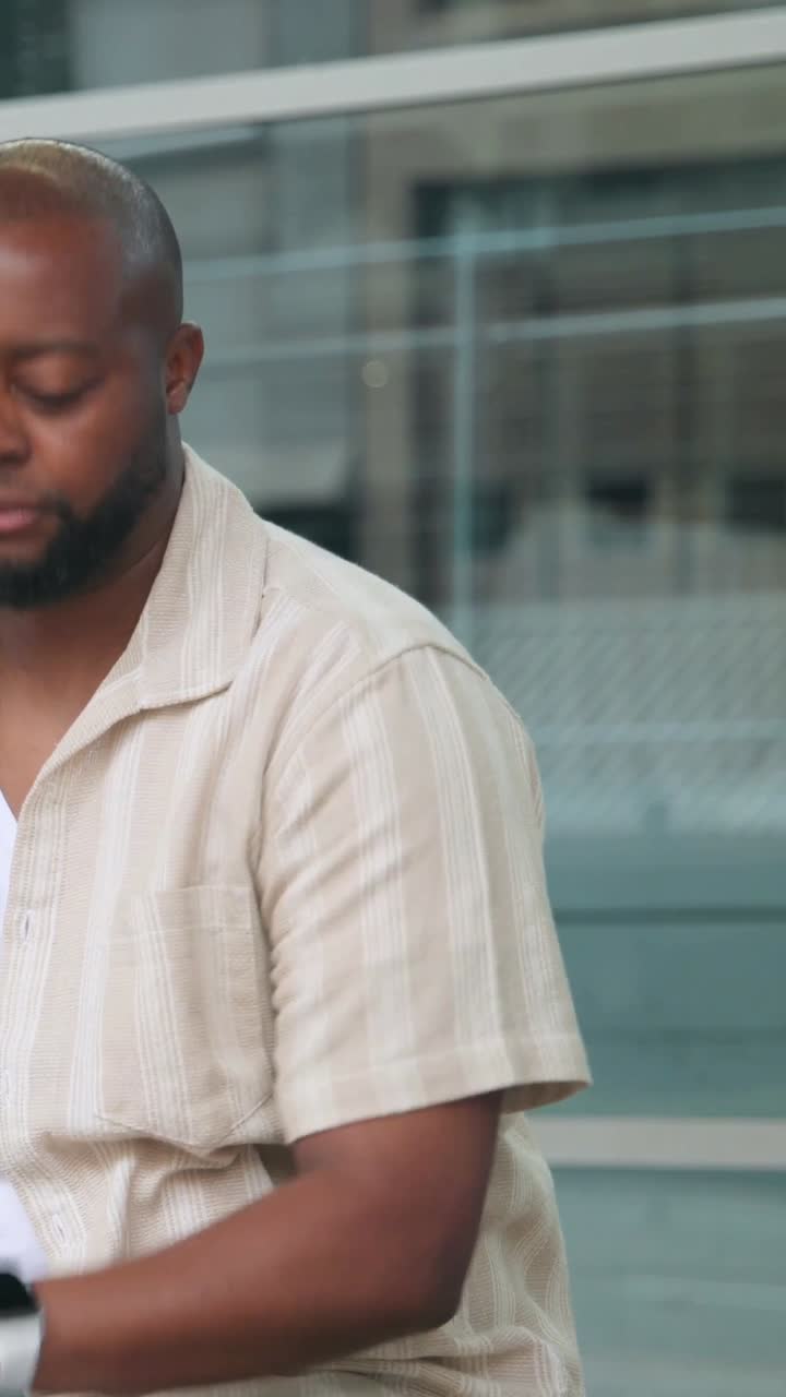 Vertical video: Looking left, man reading phone texts on bench by glass facade, shirt, copy space
