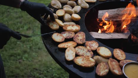 Chef-Masculino-Preparando-Papas-Y-Verduras-Para-Misiones-En-Una-Parrilla-Abierta-En-El-Patio-Trasero-En-La-Noche-De-Verano-Con-Guantes-Negros-En-Primer-Plano