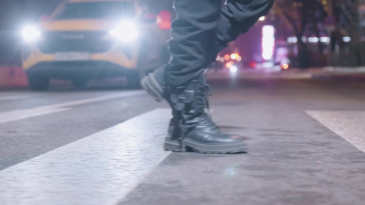 Night city crosswalk combat boots stride, lone pedestrian moves across zebra under harsh headlights, wet asphalt reflecting neon, heavy laced boots scuffing paint, cinematic tension and urban