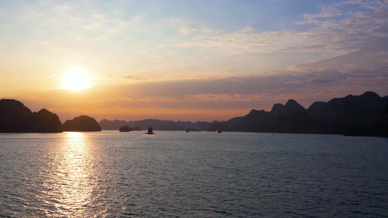 A captivating sunset illuminates Ha Long Bay's mystical seascape, where towering limestone formations create silhouettes against a golden sky as boats navigate the peaceful waters