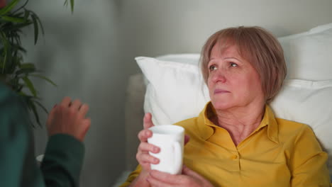 sad senior woman takes cup of water resting in bed at home