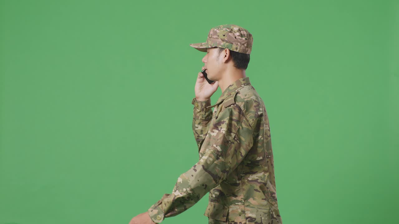 녹색 스크린 배경 스튜디오에서 는 동안 스마트폰으로 이야기하고 주변을 둘러보는 아시아 남성 군인의 측면 시각