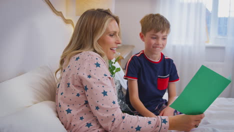 hijo amoroso sorprendiendo a la madre en la cama con flores y tarjeta casera en el cumpleaños o el día de la madre