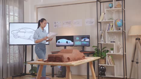 asian woman automotive designer looking at car design sketch papers in her hands and walking around in the studio with the sculpture of car clay