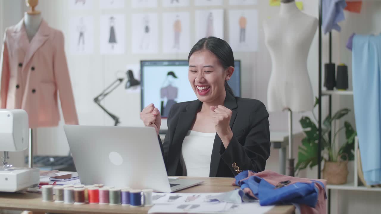 사업복을 입은 행복한 아시아 여성 디자이너가 노트북에서 일하고 스튜디오에서 옷을 디자인하는 동안 축하하고 있습니다.