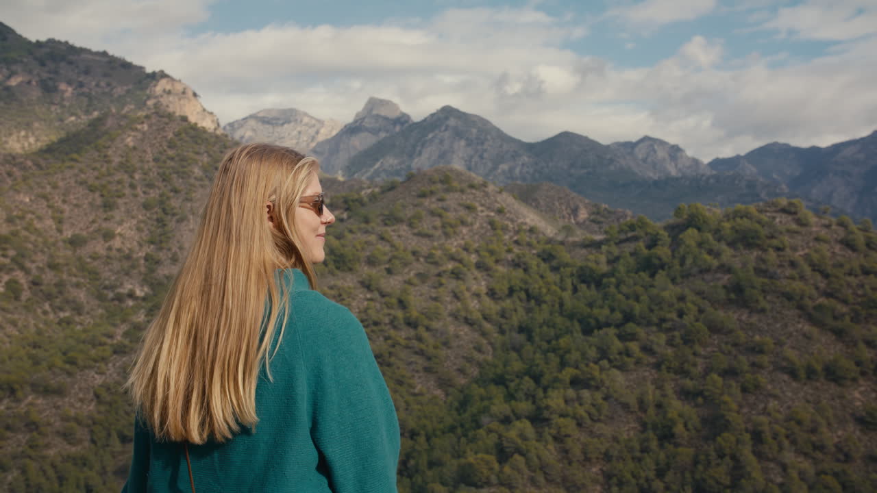 A beautiful blonde woman with long blonde hair stands facing a scenic mountain landscape in Southern Spain. Captured from behind, she gazes into the vast, rugged terrain