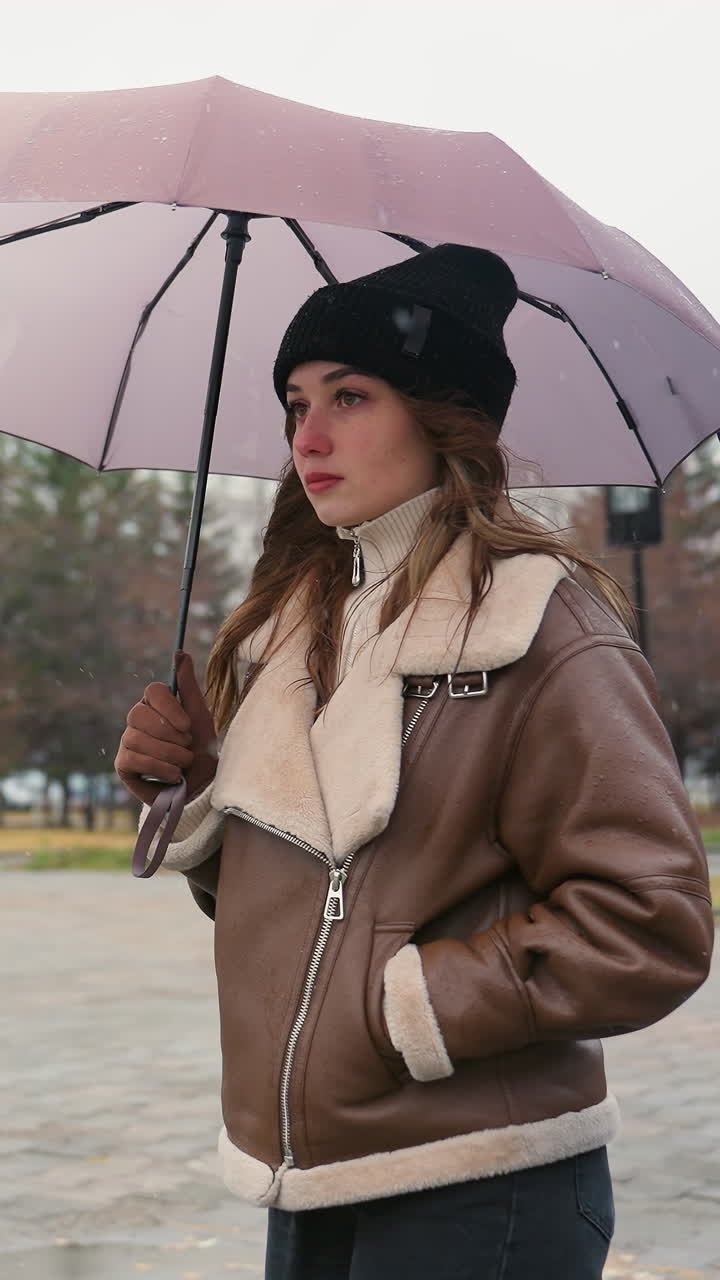 Ragazza pensierosa che tiene un ombrello, indossa un berretto di lana nero e una giacca shearling marrone, circondata da pini ed edifici urbani, con una leggera nevicata in una fredda giornata nuvolosa