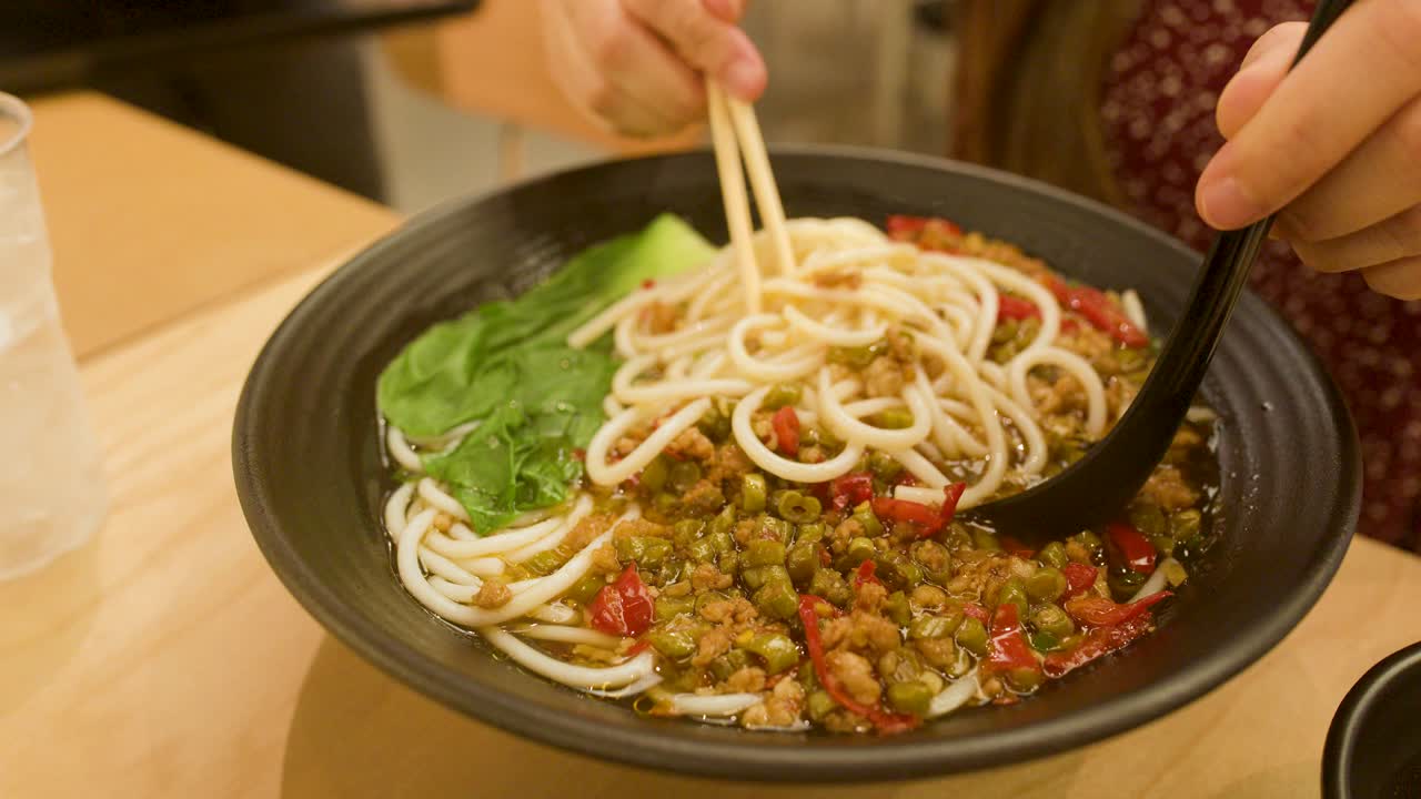 Person uses chopsticks and spoon to mix spicy noodle soup with vegetables and minced meat