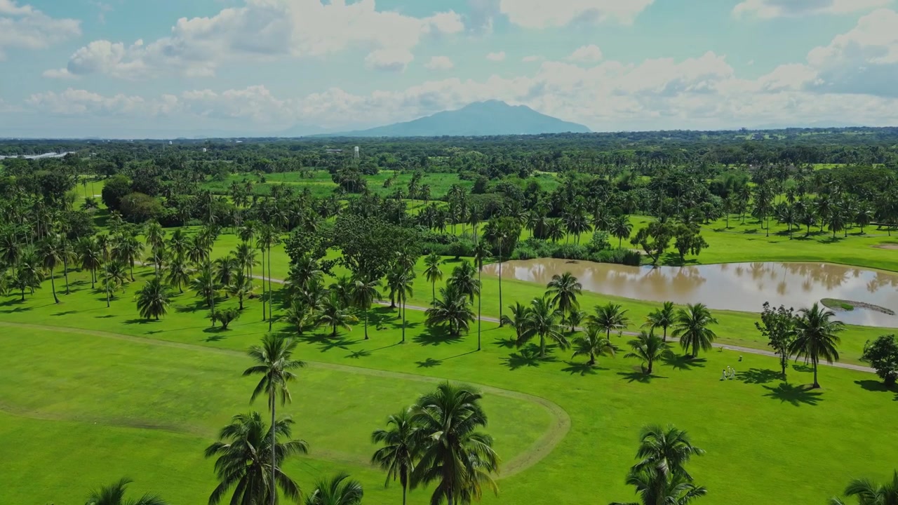 Wide aerial shot entering Canlubang Golf and Country Club in Calamba Laguna Philippines showcasing a luxury country club surrounded by lush green fairways, towering palm trees during bright daylight.
