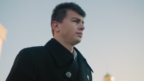 confident man in black coat and white shirt focuses on his phone outdoors under a soft sky, subtle movement of returning phone to pocket reflects calm determination in urban corporate setting