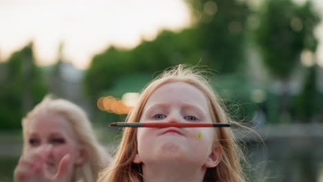 close up of fair skinned girl balancing pencil on nose with playful stare at sunset park, blurred adult figure smiling in background, colorful paint dots on cheek creating whimsical artistic mood