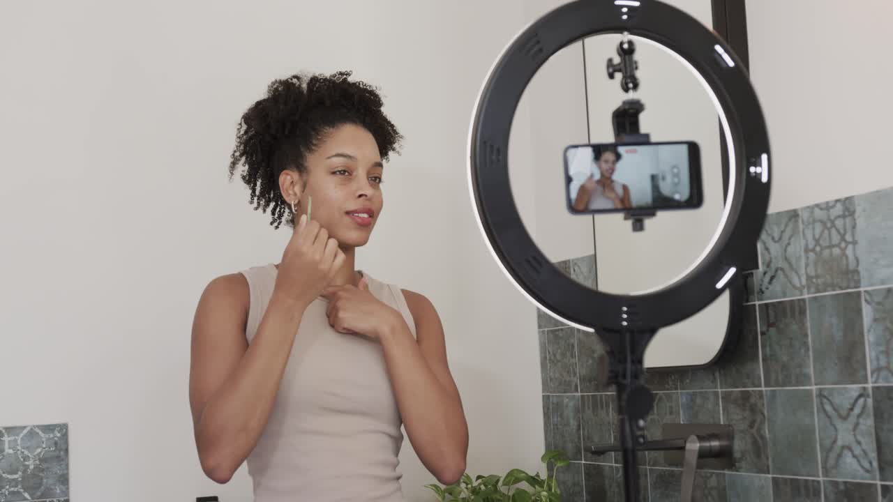 African American woman adjusting earring, rubbing jaw at sink, showing jade tool for skin on phone