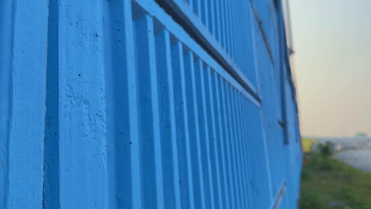 Tracking shot beside a blue-painted concrete overbridge wall, revealing texture and depth as vehicles pass on the nearby road under soft morning light