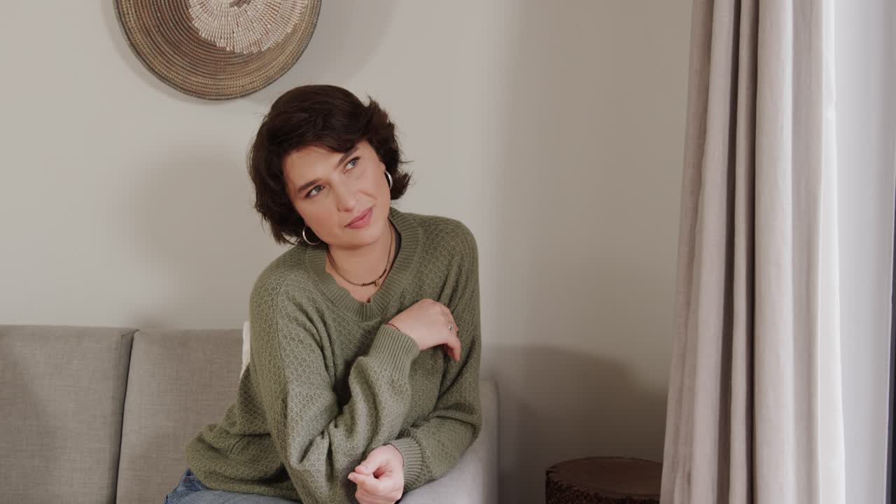 Woman settling into gray sofa, thinking, tilting head, smiling and laughing while warming to camera